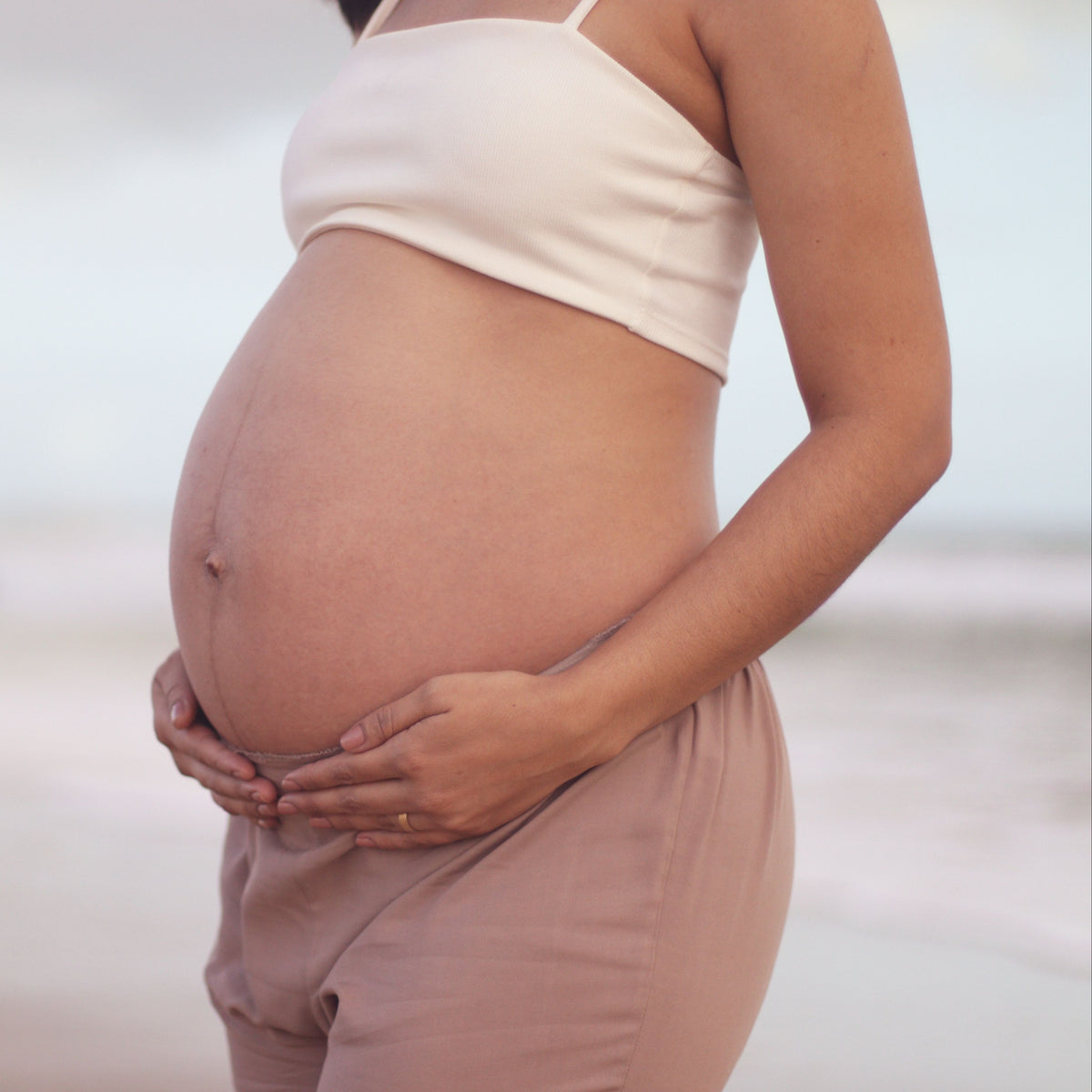 Pregnant person wearing a beige top and light pink pants, standing against a blurred background.