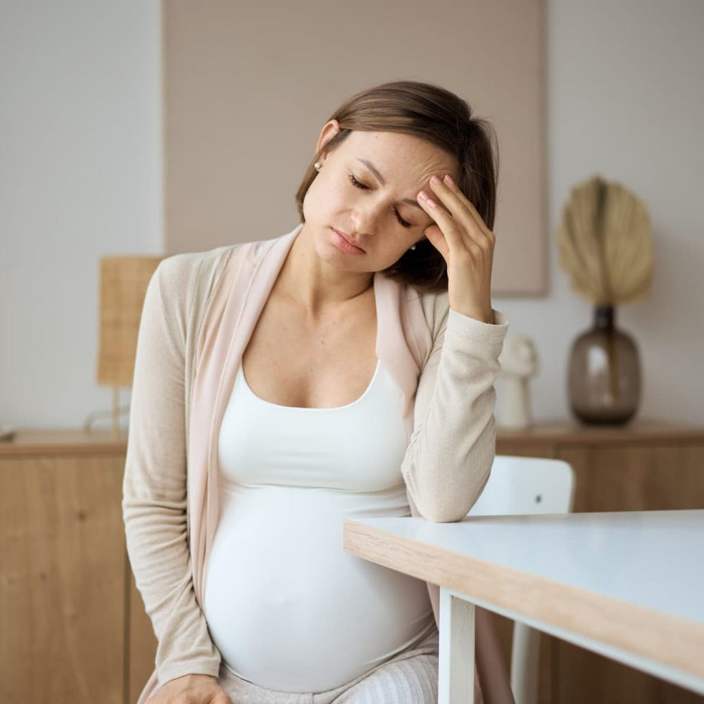 A pregnant woman in a white tank top and beige cardigan sits at a table in a softly lit room, holding her head in her hand with a look of fatigue or pain.